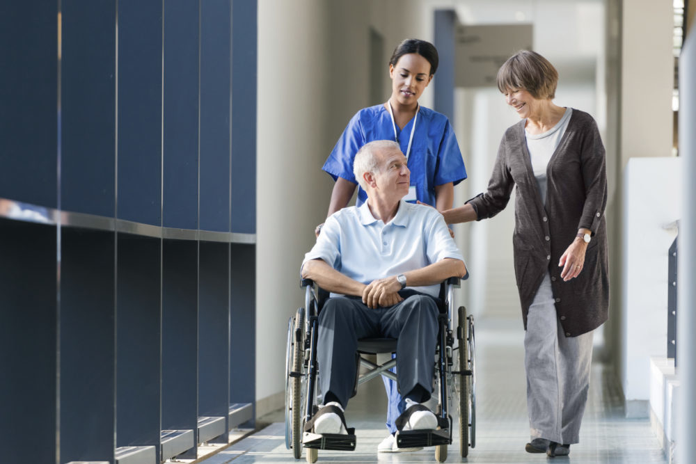 Elderly man in a wheelchair being supported by older woman and clinical staff member